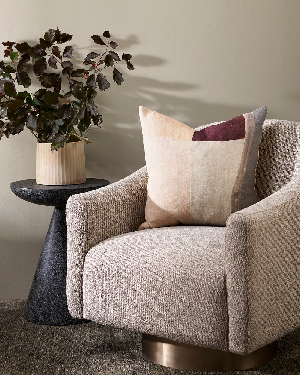 benny cushion from baya with grey, port red, beige and cream brushstrock pattern on a grey boucle fabric chair in cream. Next to it is a round coffee table in dark grey stone with a cream ridged pot with greenery in it