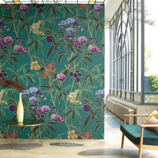 Camelia wall panel by James Dunlop from Casadeco. Teal background with vivid plum, purple, teal and green foliage. On wall in a room with grey concrete floor, teal and light wood sofa, small round gold side table with elongated cream vase on. Windows to the right are arched with grey frames