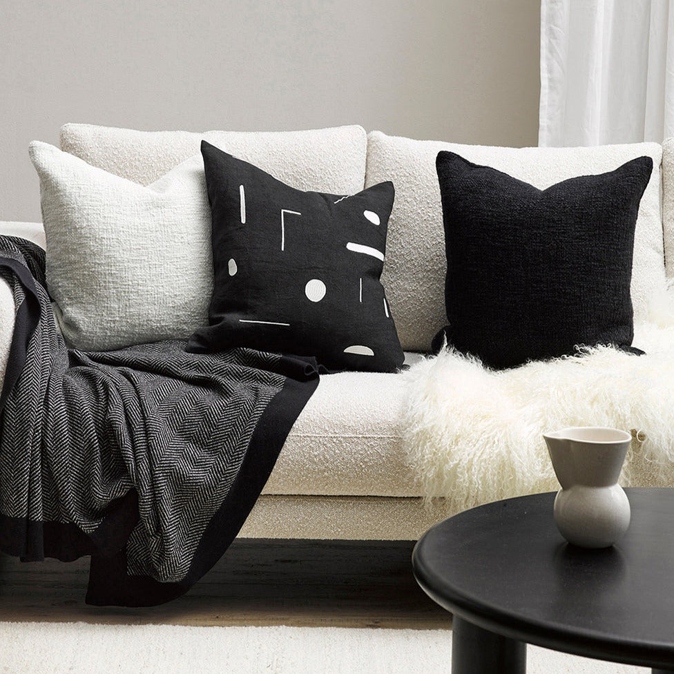 Organic cotton Carmel throw in peat on a white boucle sofa with two other cushions, one white and one black with white spots and lines, also a tibetan lambskin throw - fluffy cream throw with a black coffee table and cream jug. Scene is set on a brown wooden flor with a ivory plain carpet