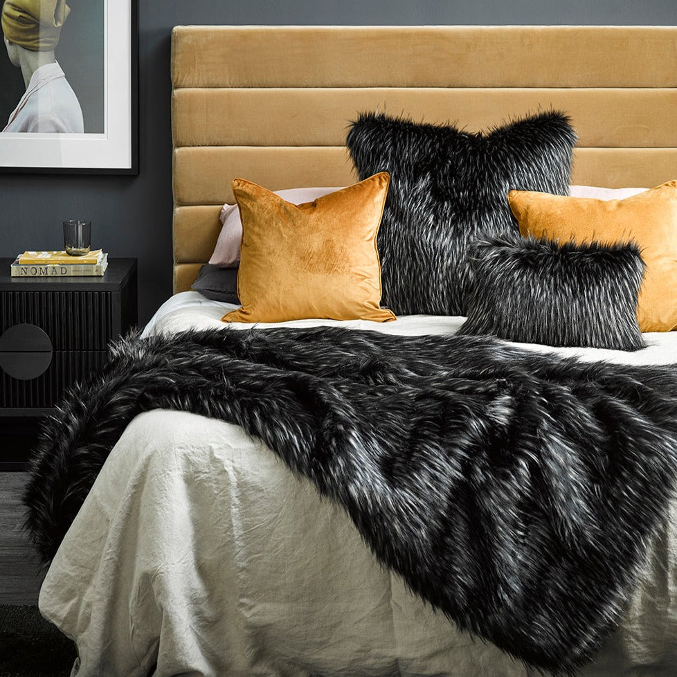 Ebony Plume imitation fur throw on bed with mustard padded headboard and cushions and a painting on grey wall and besdie table