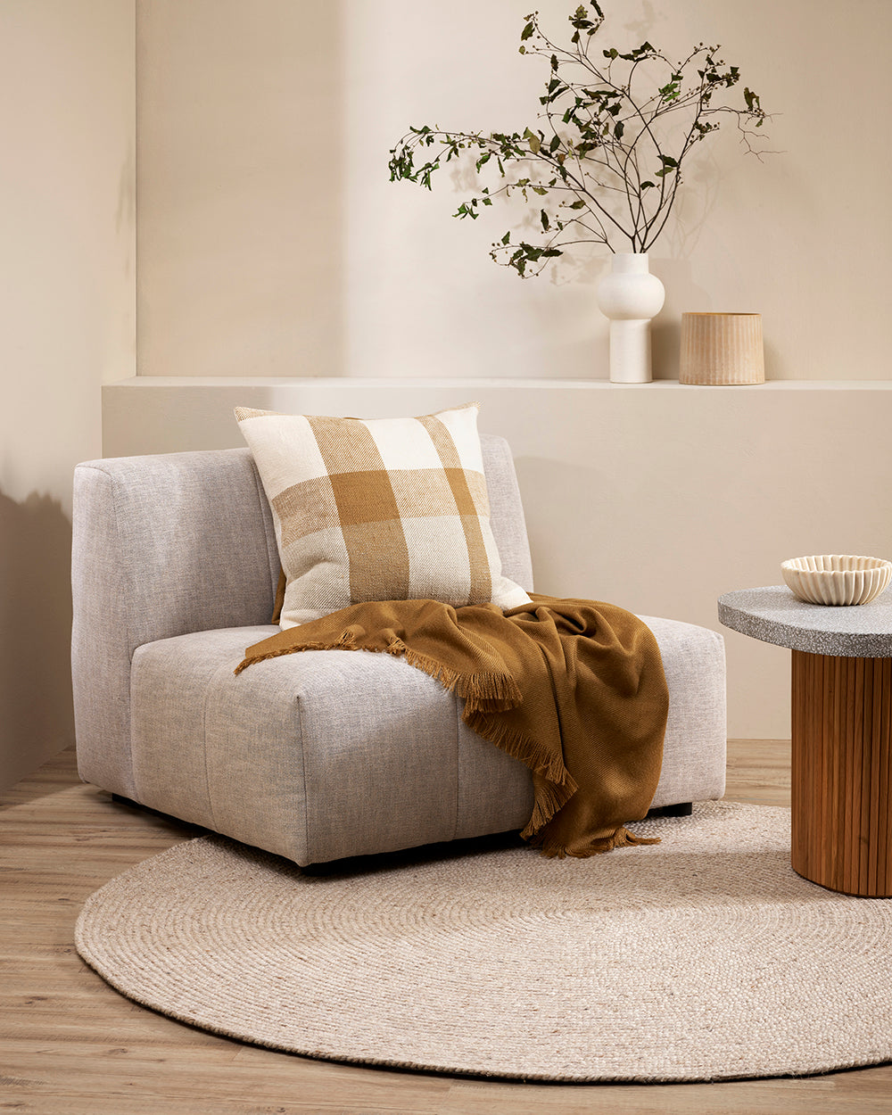 Jefferson cushion cumin on a cream sofa chair with a cumin coloured throw. The room features a low wall behind the sofa chair with a white vase and greenery and a beige coloured smaller vase. Ther eis a round sisal type rug on a wooden floor with a wooden side table and concrete top with a cream ridged bowl on top