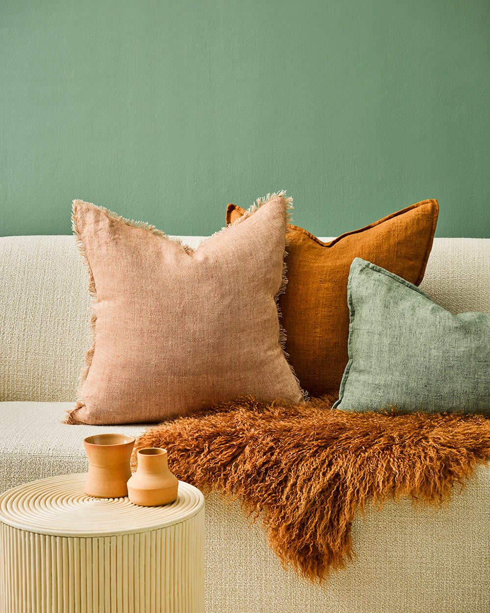 Keaton Linen cushion in cinnamon with tobacco and great cushions and lamb fur throw on cream couch with sage green background