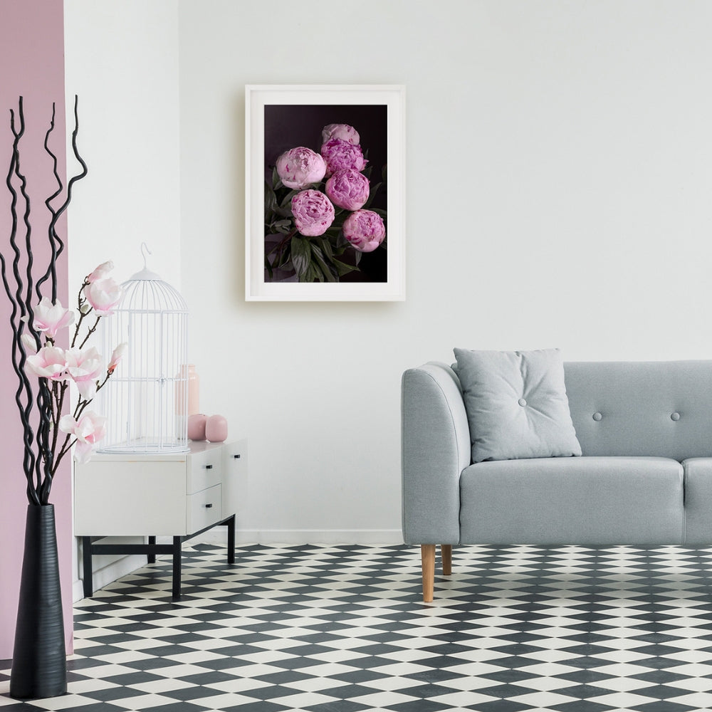 bunch of pink peonies on a black back ground on the wall wth a grey sofa, grey and white diamond check floor, white side table with pink candle and vases with a white bird case