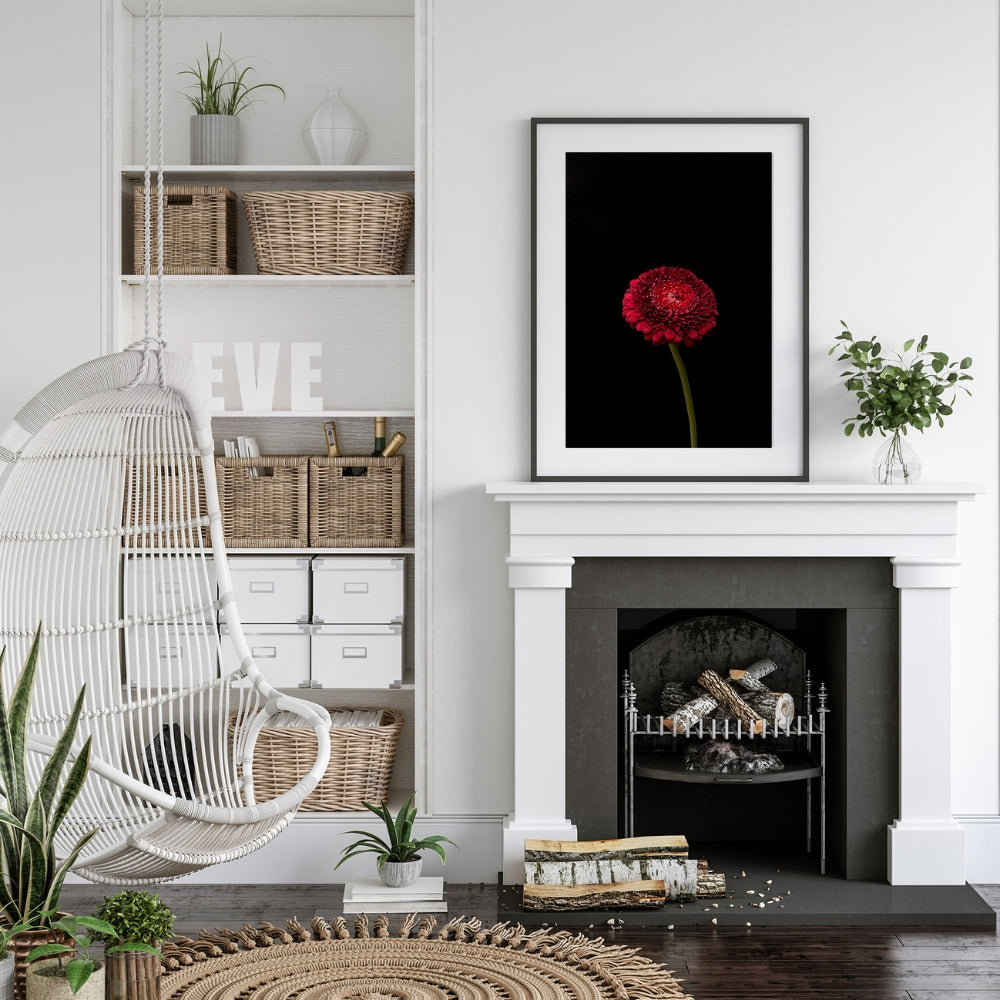 single stem red gerbera floral art picture in a white room above a white fire place with a hanging white cane chair, white bookcase with wicker baskets and vases. Brown wooden floor with circular rattan rug