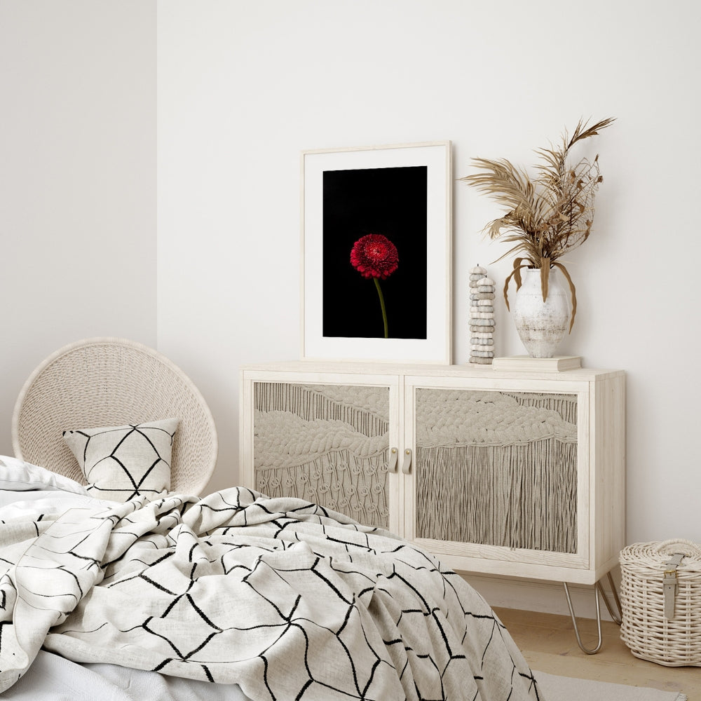 red gerbera picture above a white side board in a white bedroom with a bed andn black and white quilt cover, same on cushions on a white wicker chair, white rattan basket on the floor