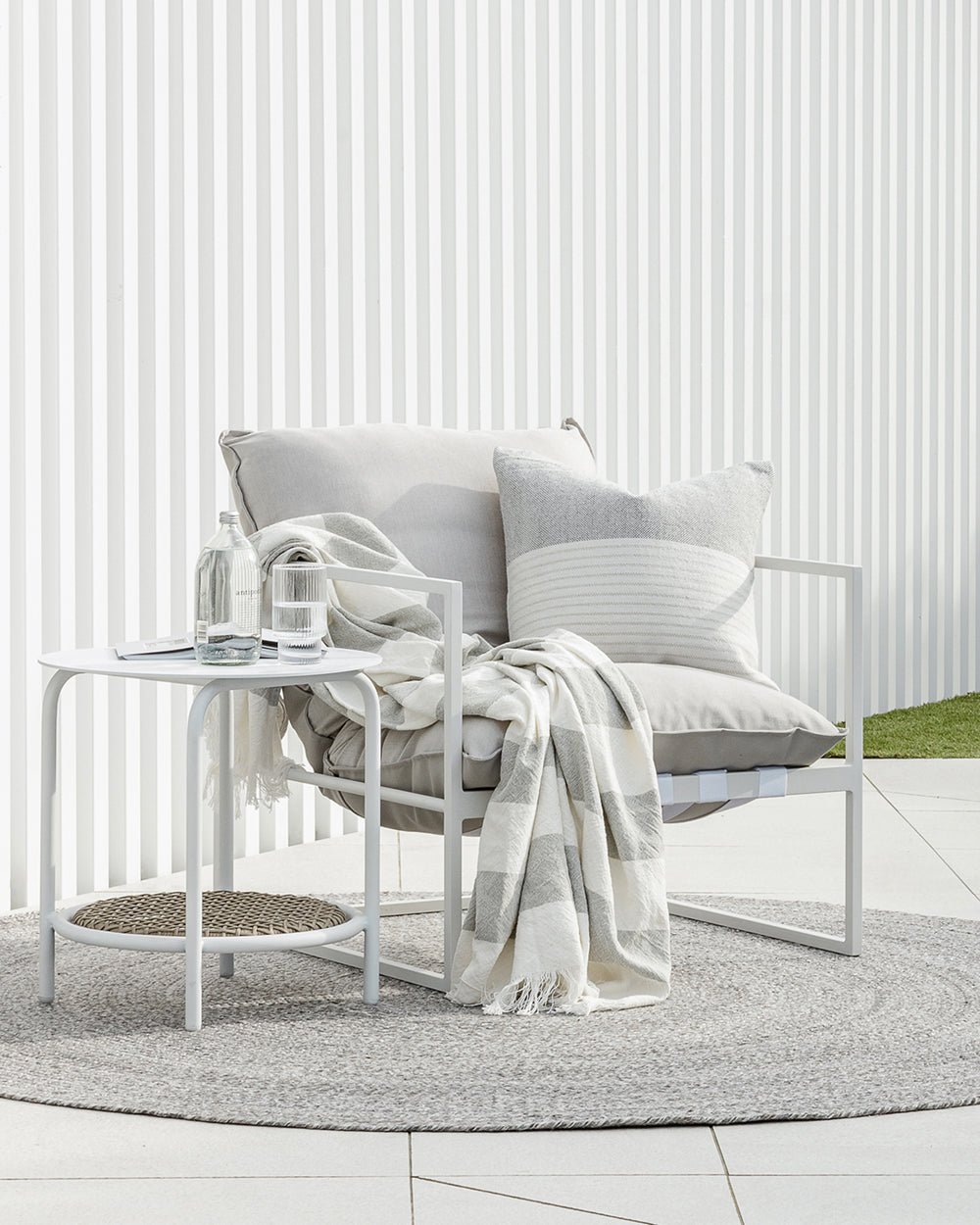 Willett outdoor cushion on patio chair with a grey and cream throw on a round mat next to a side table witih water and glass